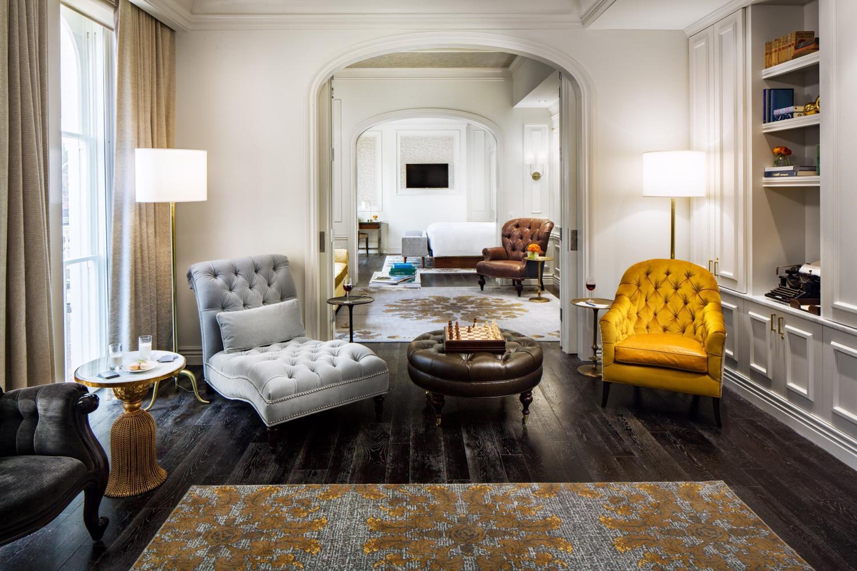 A hotel room with a tufted armchair, a yellow armchair, a grey tufted sofa, a wooden coffee table, and a patterned rug.