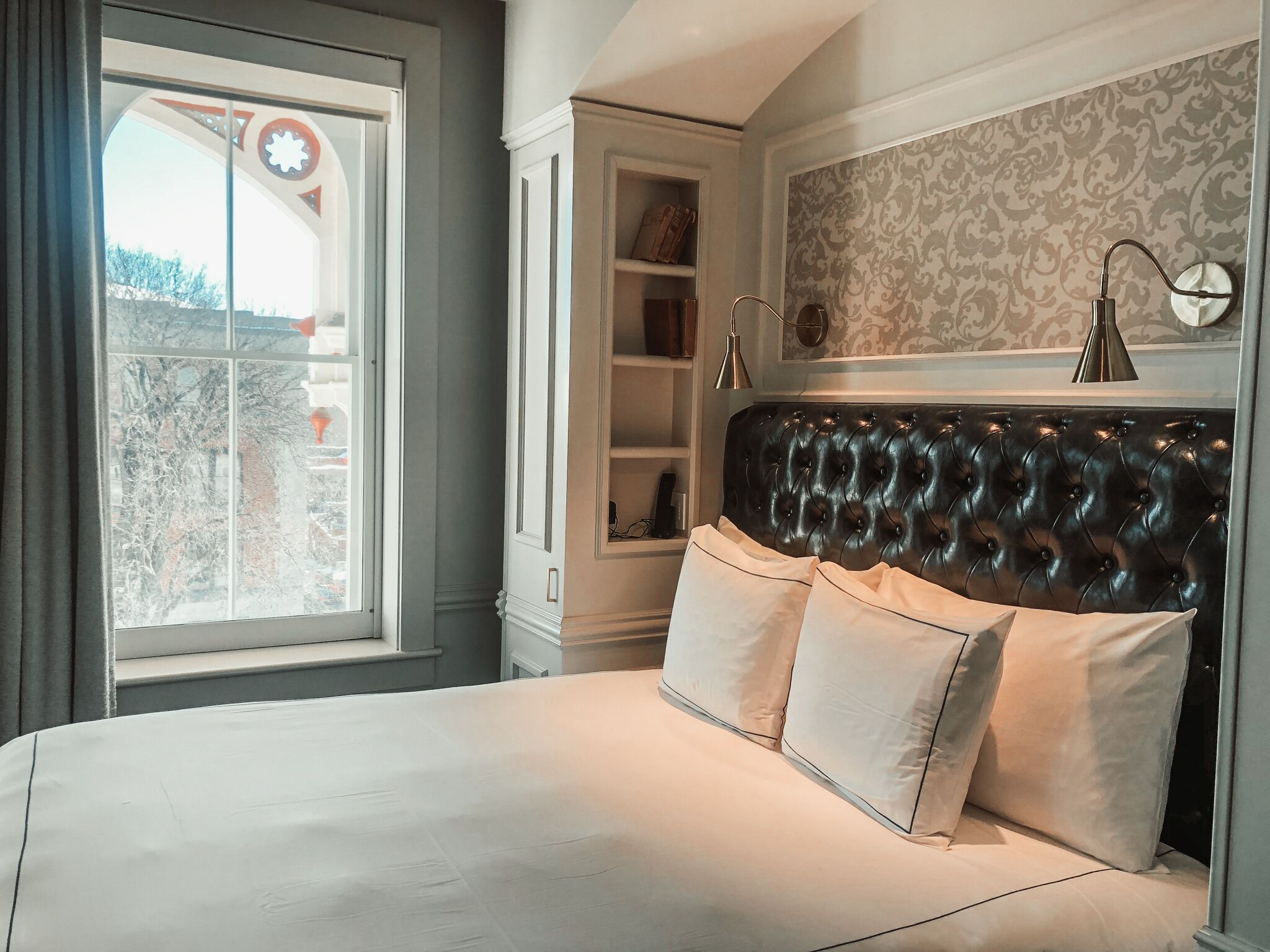 Une chambre à coucher avec un grand lit, une tête de lit, une fenêtre, une bibliothèque et un mur recouvert d’un papier peint à motifs.