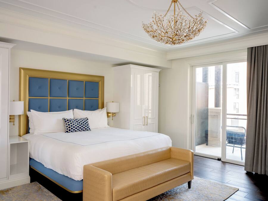 Ein Hotelzimmer mit einem großen Bett, einem goldfarben gerahmten Kopfteil, einer Bank, einer Kandelaber und Blick ins Freie.