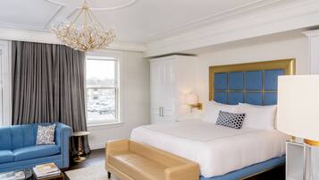 A hotel room with a large bed, a blue sofa, a beige armchair, a chandelier, and a view of the city through the window.