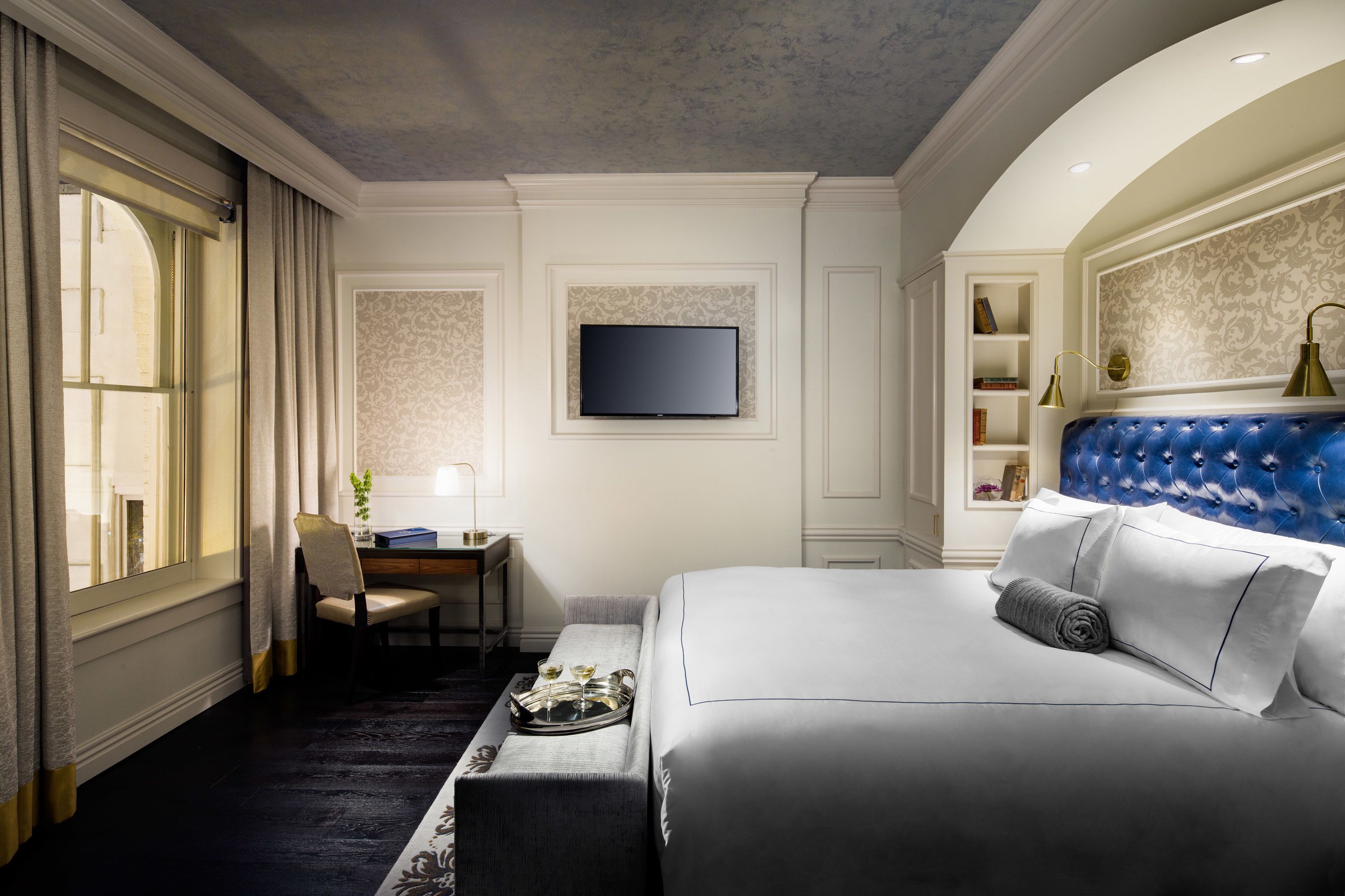 A hotel room with a large bed, a desk with a chair, a television mounted on the wall, and a bookshelf.