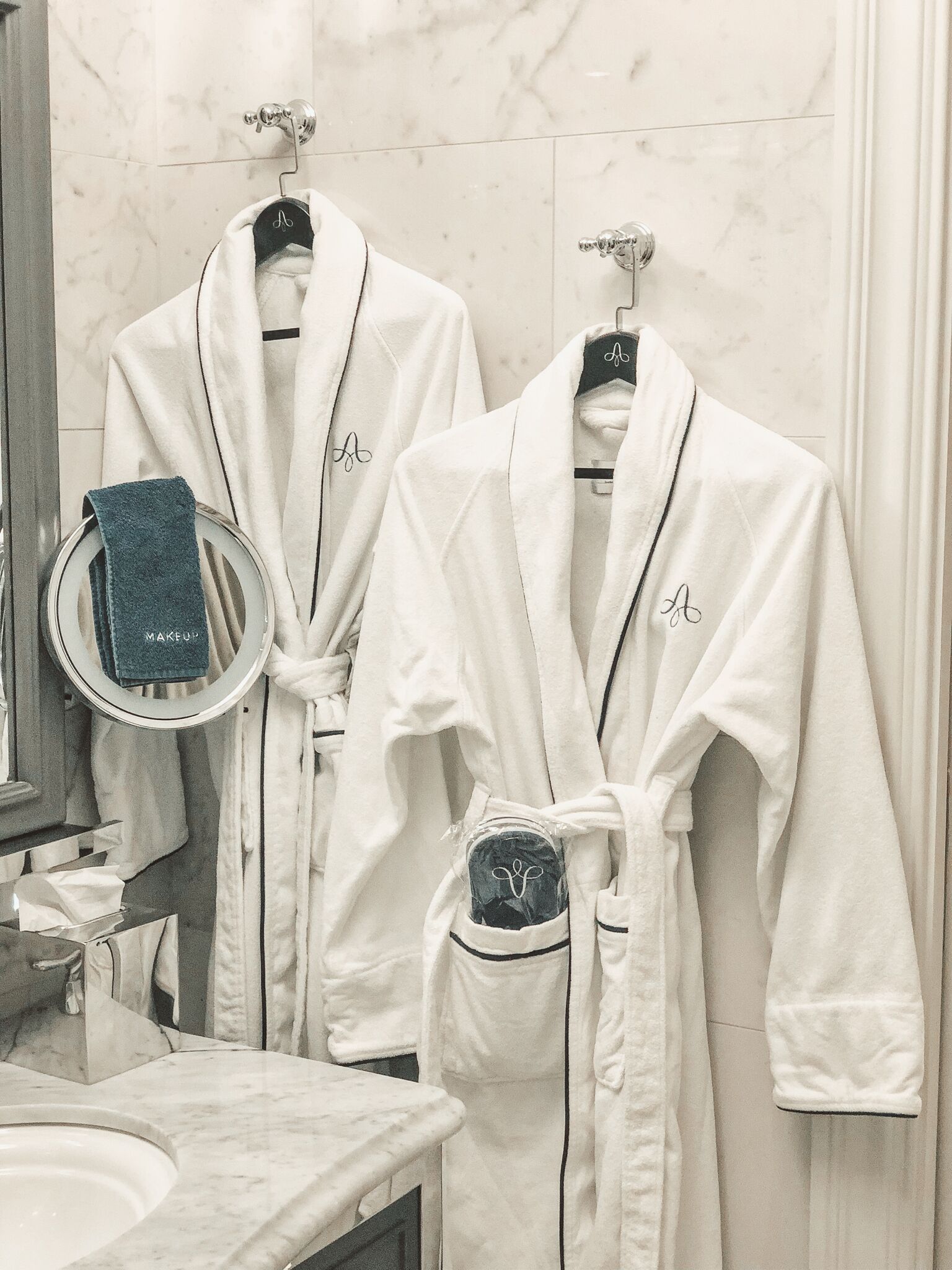 Un bagno con due accappatoi bianchi appesi a dei ganci, uno specchio tondo e un asciugamano blu su un portascopetti circolare.