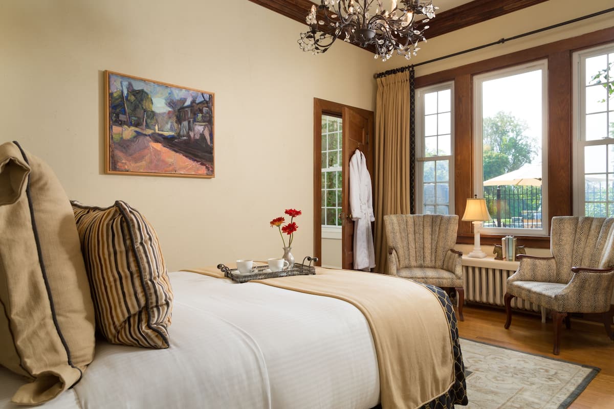 A hotel room with a large bed, two armchairs, a small table with a lamp, a painting on the wall, and a window with curtains.
