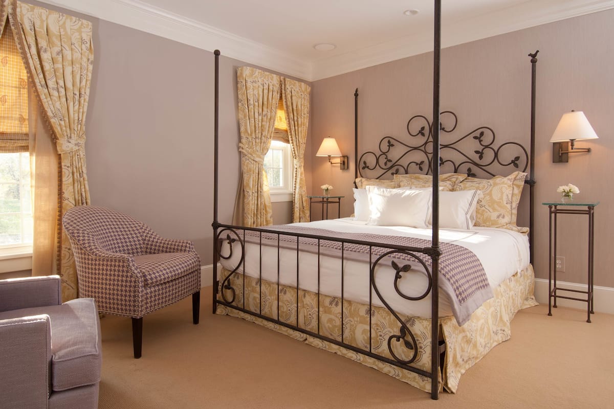 A four-poster bed with a metal frame, a houndstooth armchair, a small table with a lamp, and a window with curtains.