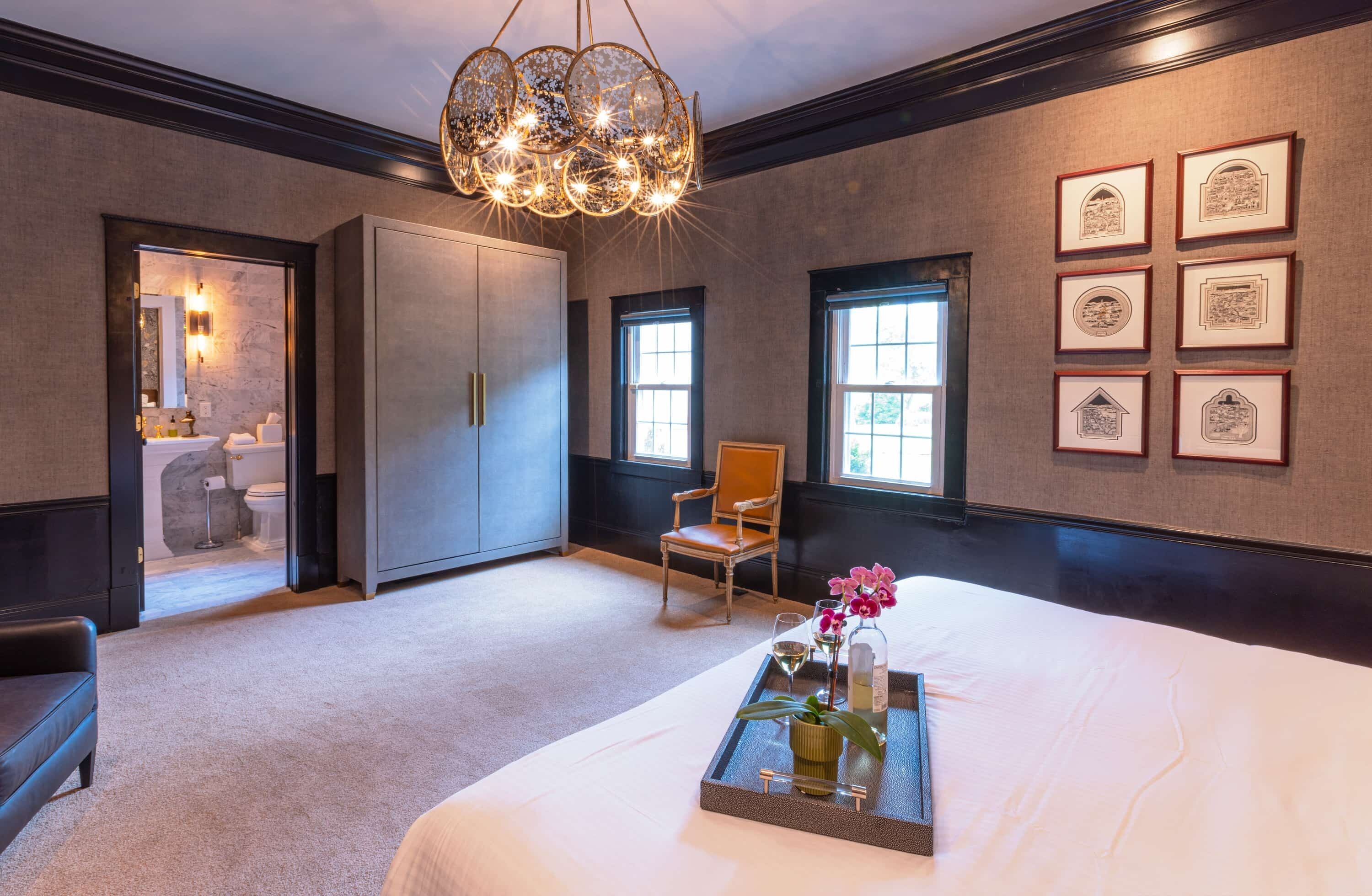 Quarto de hotel com uma cama grande, um candelabro, quadros nas paredes e uma bandeja com flores e uma garrafa sobre a cama.