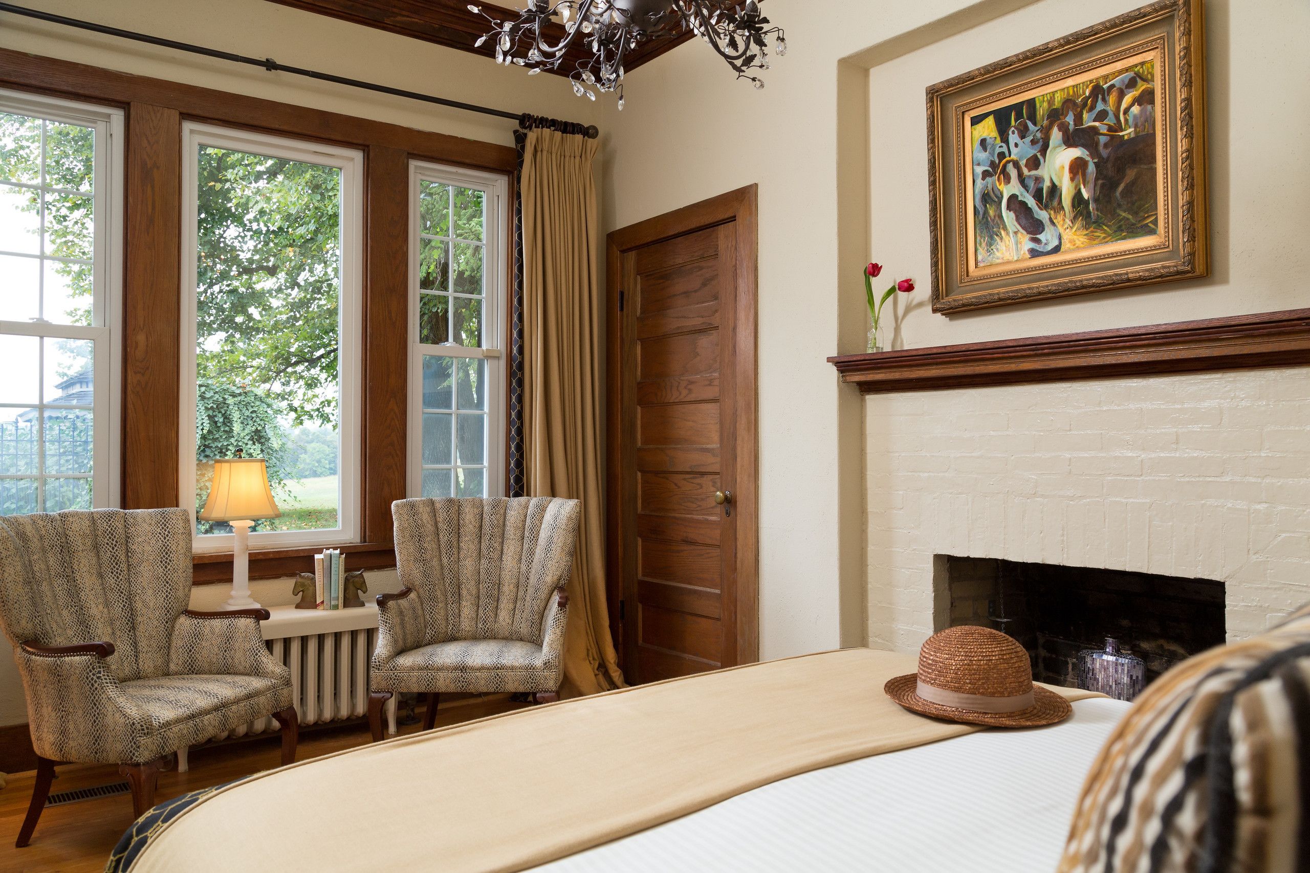 A room with a fireplace, two patterned armchairs, a bed, and a painting on the wall.