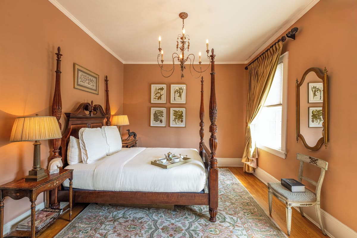 A bedroom with a four-poster bed, a chandelier, framed botanical prints, and a bench by the window.