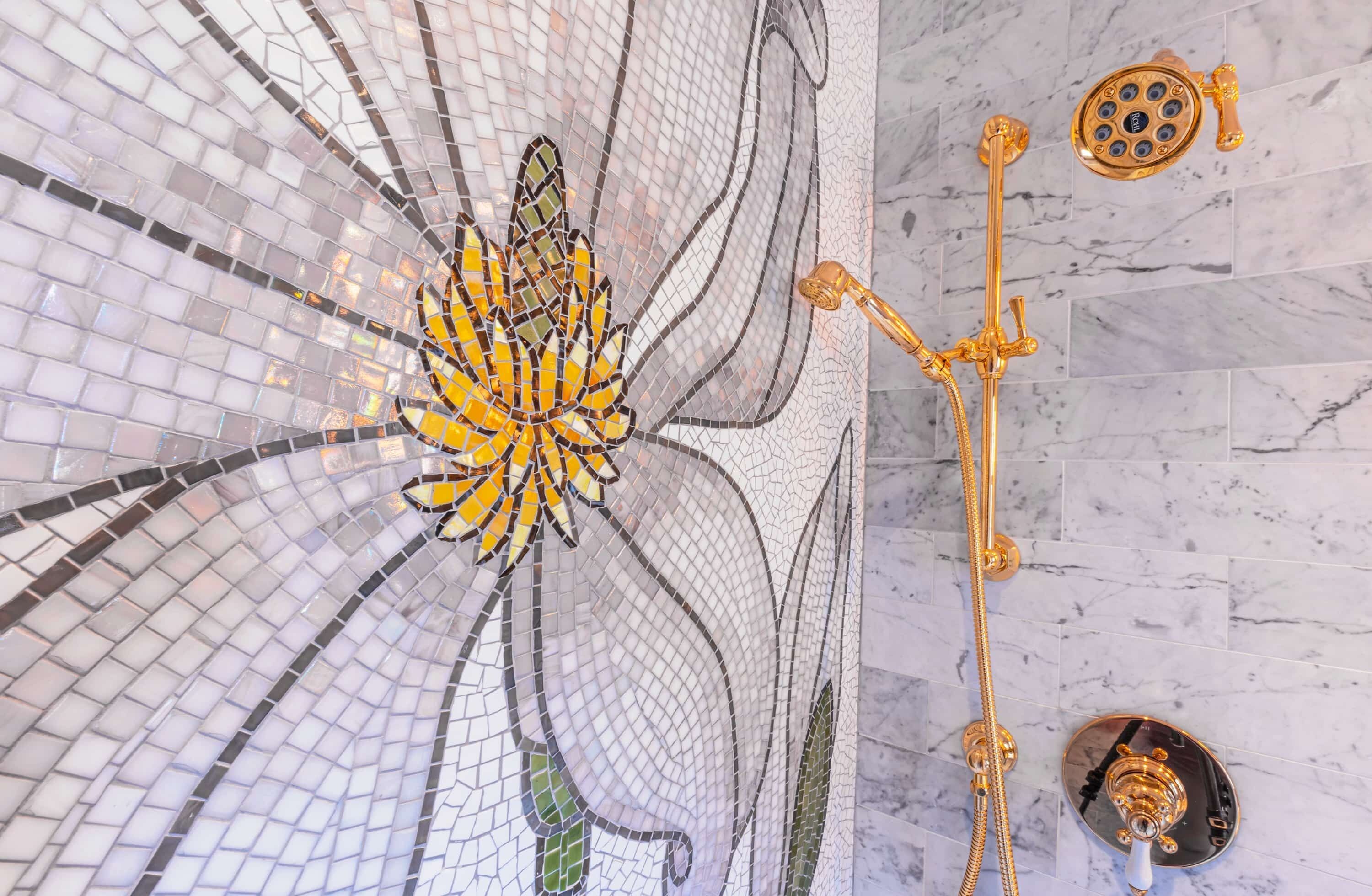 Un mosaico de una flor en una pared de azulejos, un cabezal de ducha dorado y un suelo de mármol.