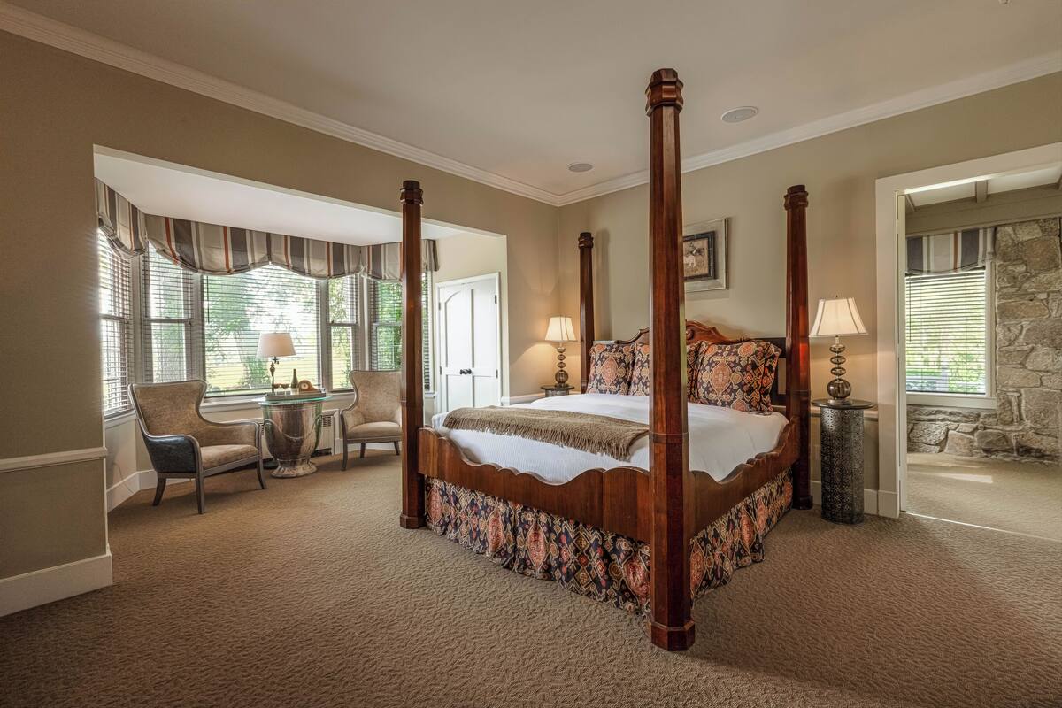 A four-poster bed with a patterned bedspread, a chair, a small table with a lamp, and a stone wall.