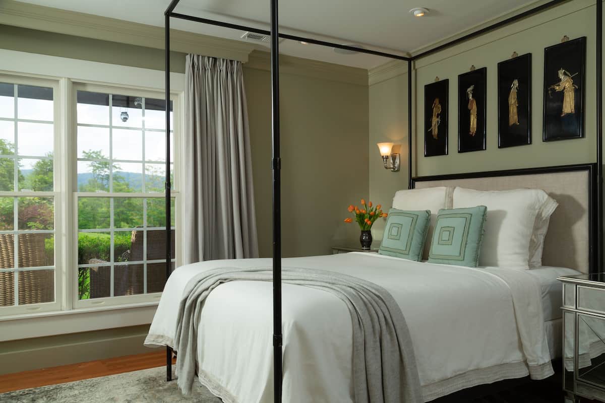 A bedroom with a large bed, a window with curtains, a nightstand with a vase of flowers, and wall-mounted artwork.