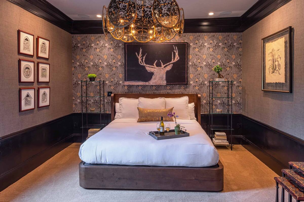 A spacious bedroom with a large bed, a chandelier, framed artwork, and a decorative plant.