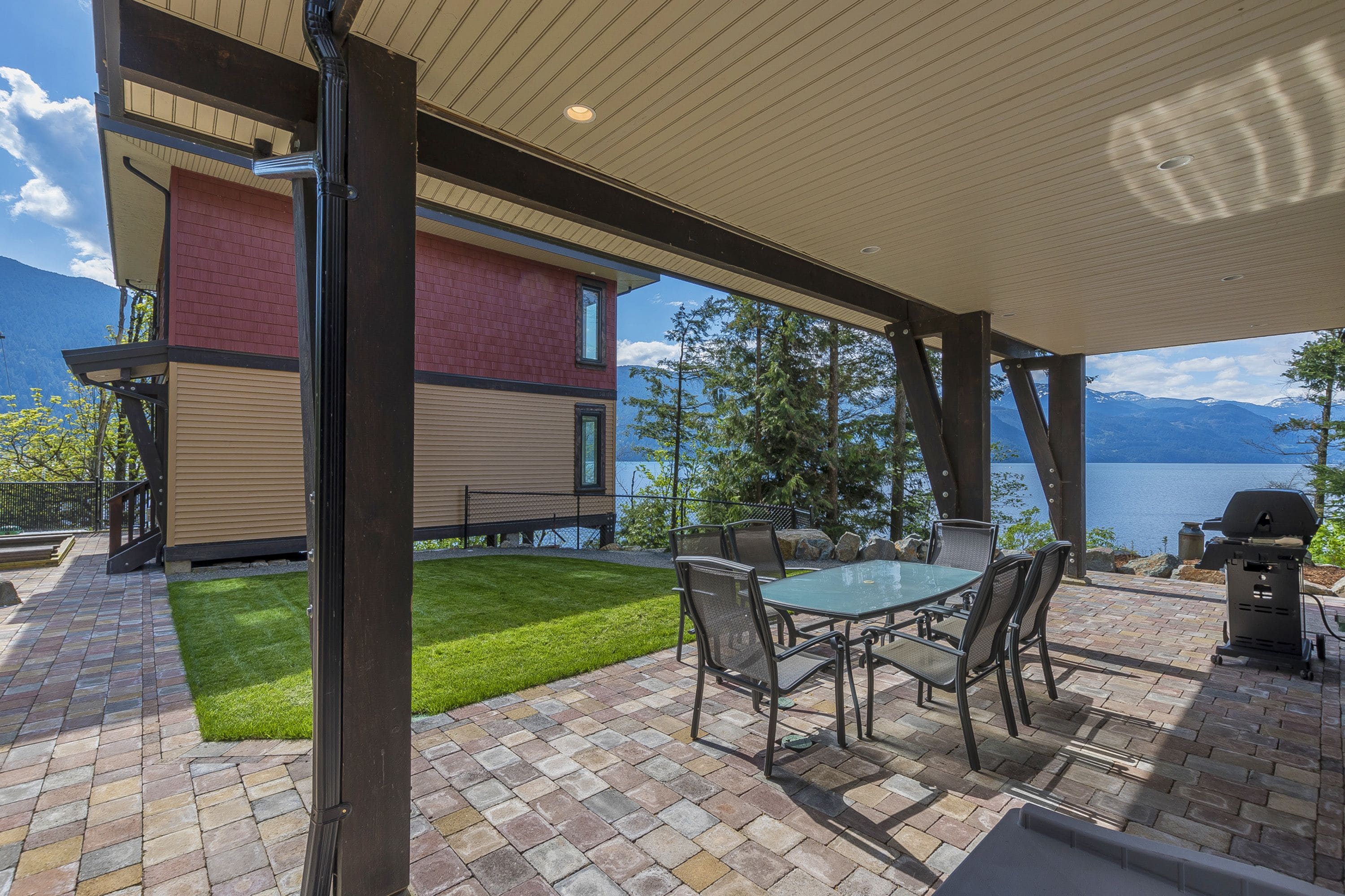 A patio with a glass table and chairs, a grill, and a view of a lake and mountains.