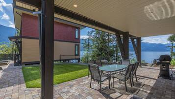 A patio with a glass table and chairs, a grill, and a view of a lake and mountains.
