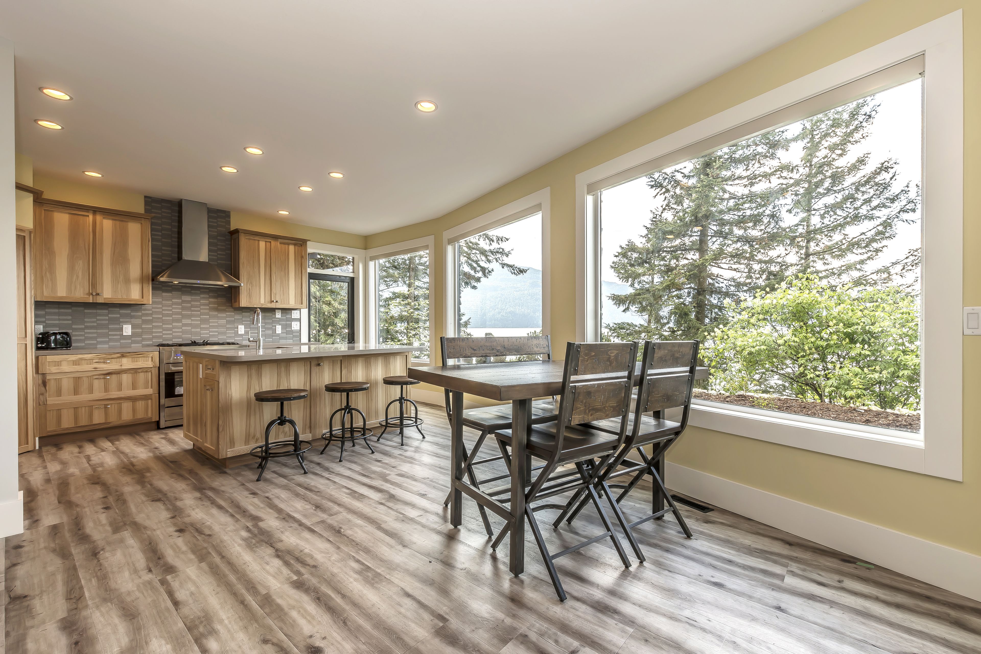 A modern kitchen with wooden cabinets, a central island with bar stools, and large windows offering a view of trees.
