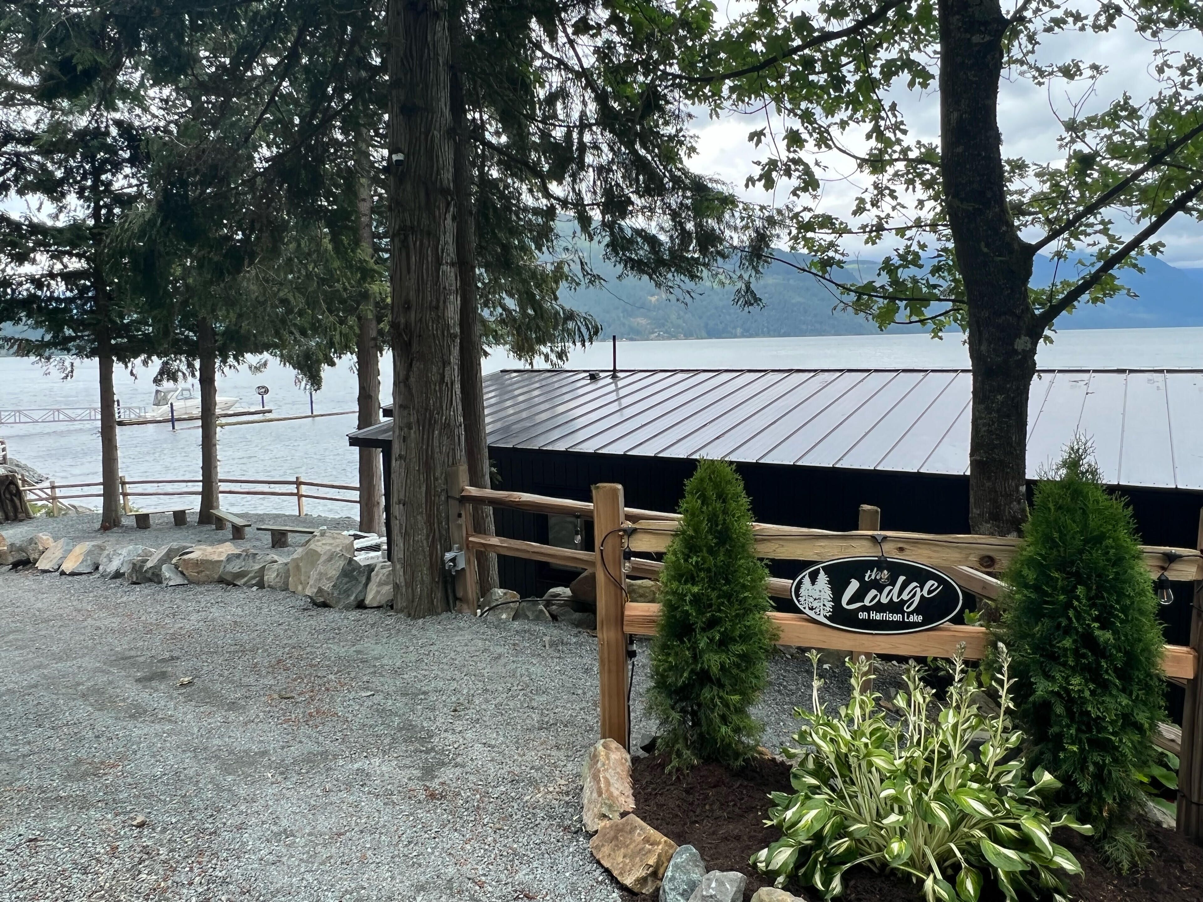 A wooden sign with 'The Lodge on Harrison Lake' text, surrounded by rocks and plants, with a view of a lake and trees in the background.