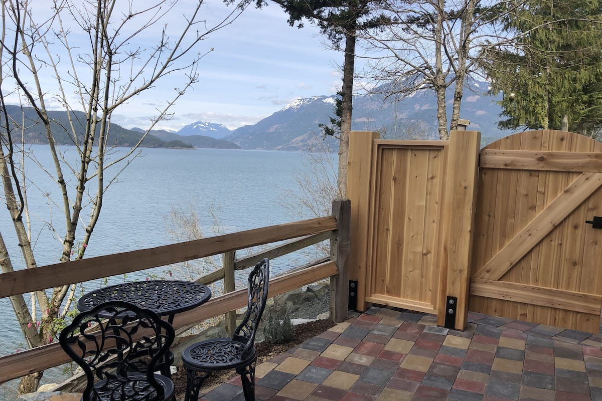 Eine Holzterrasse mit Blick auf einen See und Berge, ausgestattet mit einem Tisch, Stühlen, einem hölzernen Tor und einem gefliesten Boden.