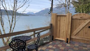 A wooden deck with a view of a lake and mountains, featuring a table and chairs, a wooden gate, and a tiled floor.