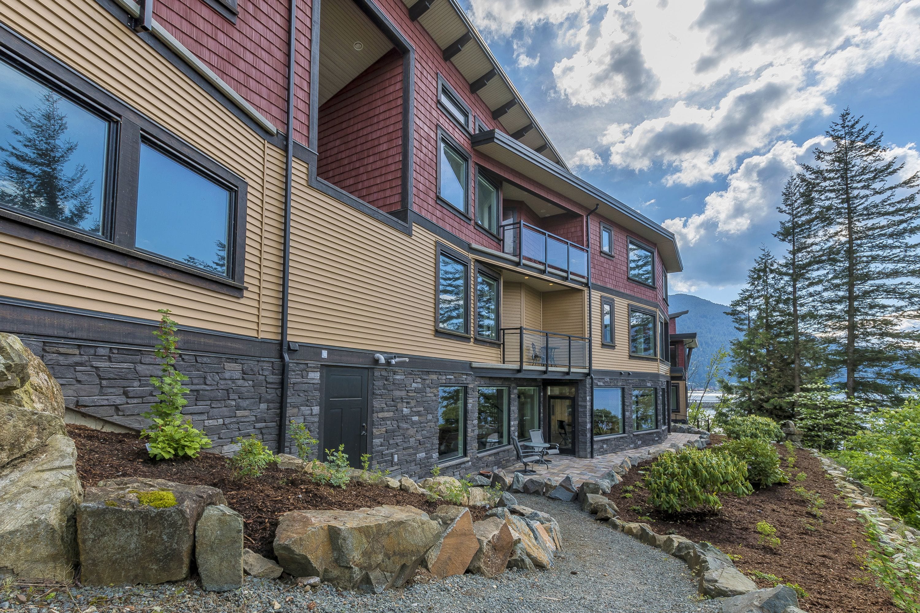 A modern multi-story house with a stone foundation, large windows, and a landscaped front yard with rocks and plants.