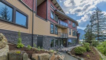 A modern multi-story house with a stone foundation, large windows, and a landscaped front yard with rocks and plants.