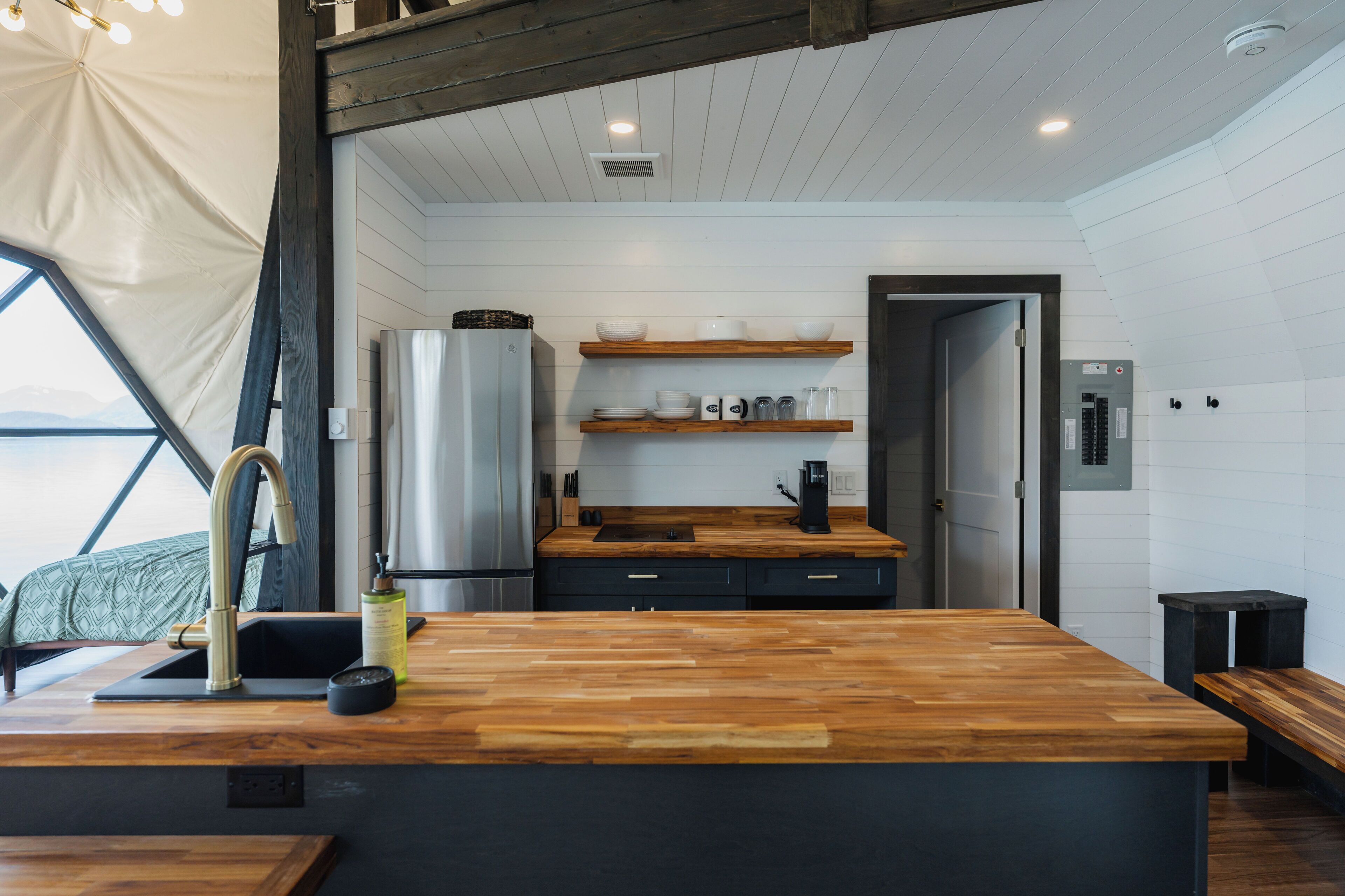 A modern kitchen with wooden countertops, a stainless steel refrigerator, and a built-in oven. There are wooden shelves above the counter and a bench to the right.