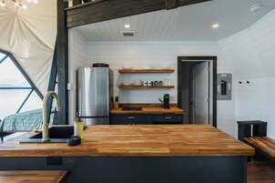 A modern kitchen with wooden countertops, a stainless steel refrigerator, and a built-in oven. There are wooden shelves above the counter and a bench to the right.