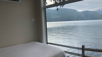 A room with a bed and a large window overlooking a lake with mountains in the background.