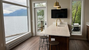 Una cocina moderna con una isla grande, un televisor montado sobre la isla y vista a un lago con montañas al fondo.
