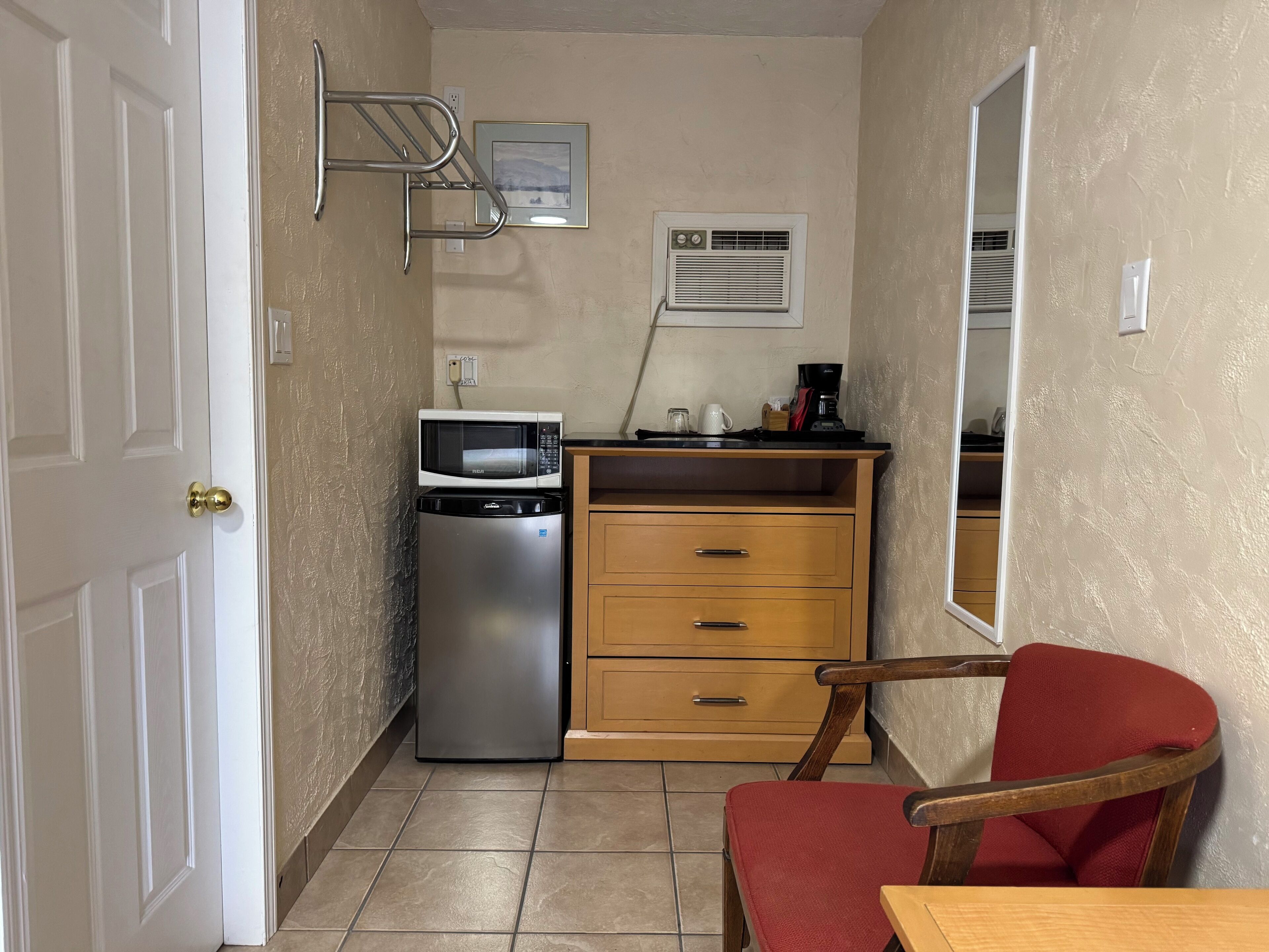 A compact kitchenette with a microwave, refrigerator, and coffee maker. There is a wooden cabinet with drawers and a red chair.