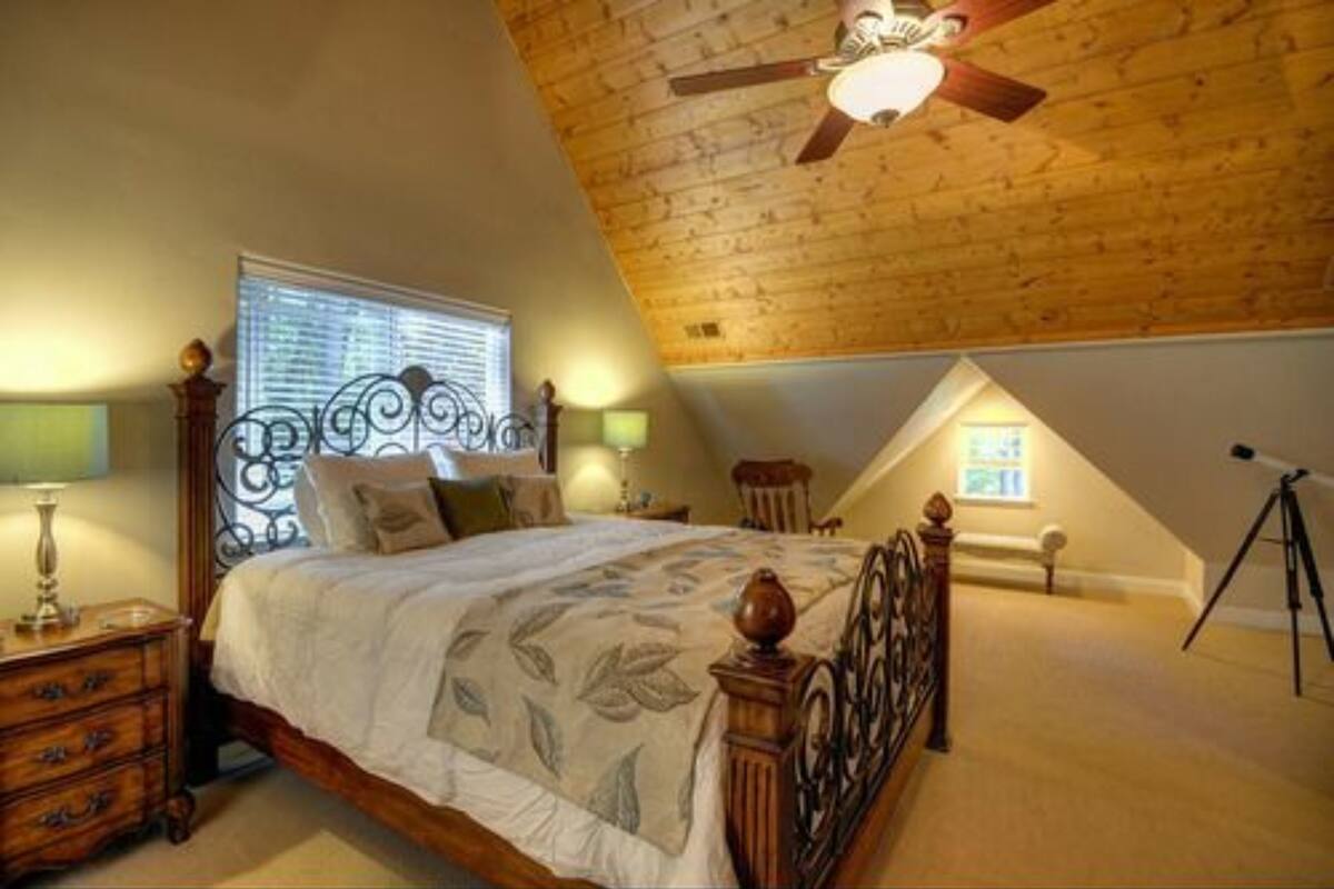 A bedroom with a wooden ceiling, a bed with a decorative headboard, a nightstand, and a window with blinds.