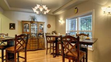 A dining area with wooden chairs and a table, a chandelier, a wooden cabinet with glass doors, and a window with a view of trees.