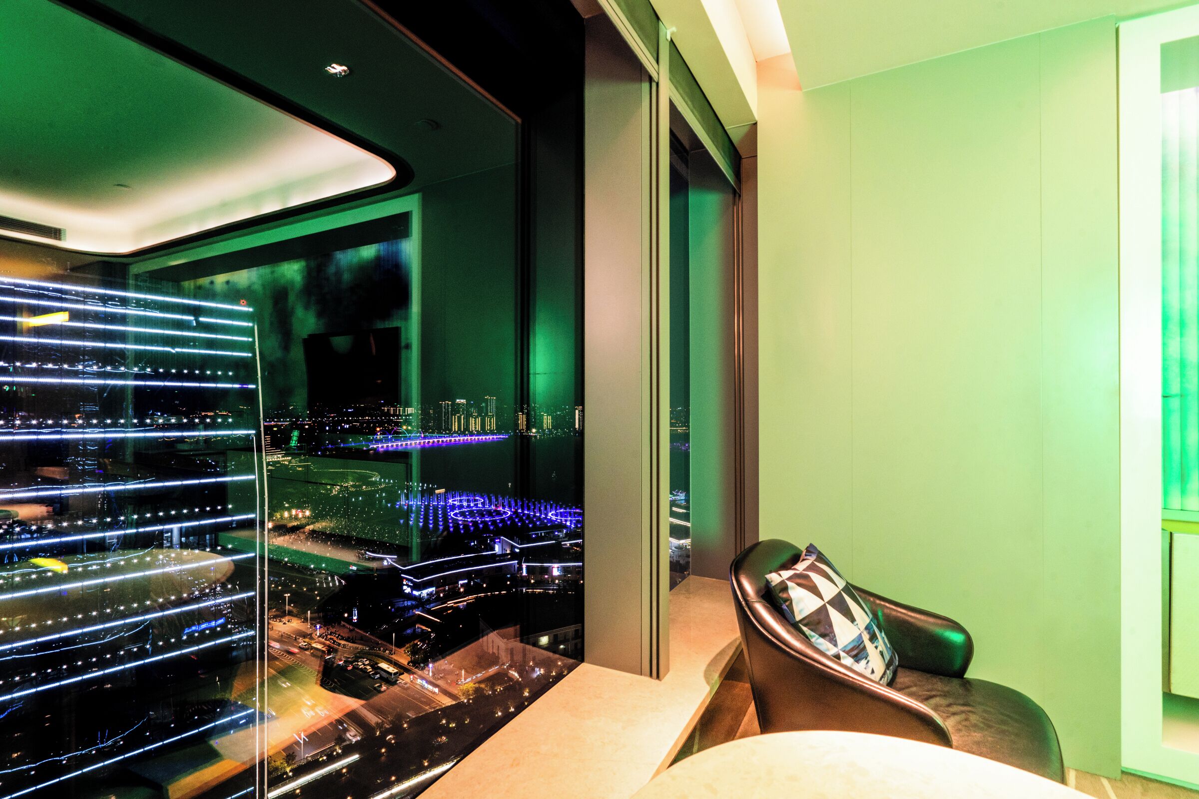 A modern hotel room with a large window offering a nighttime city view, a comfortable seating area, and a well-lit interior.