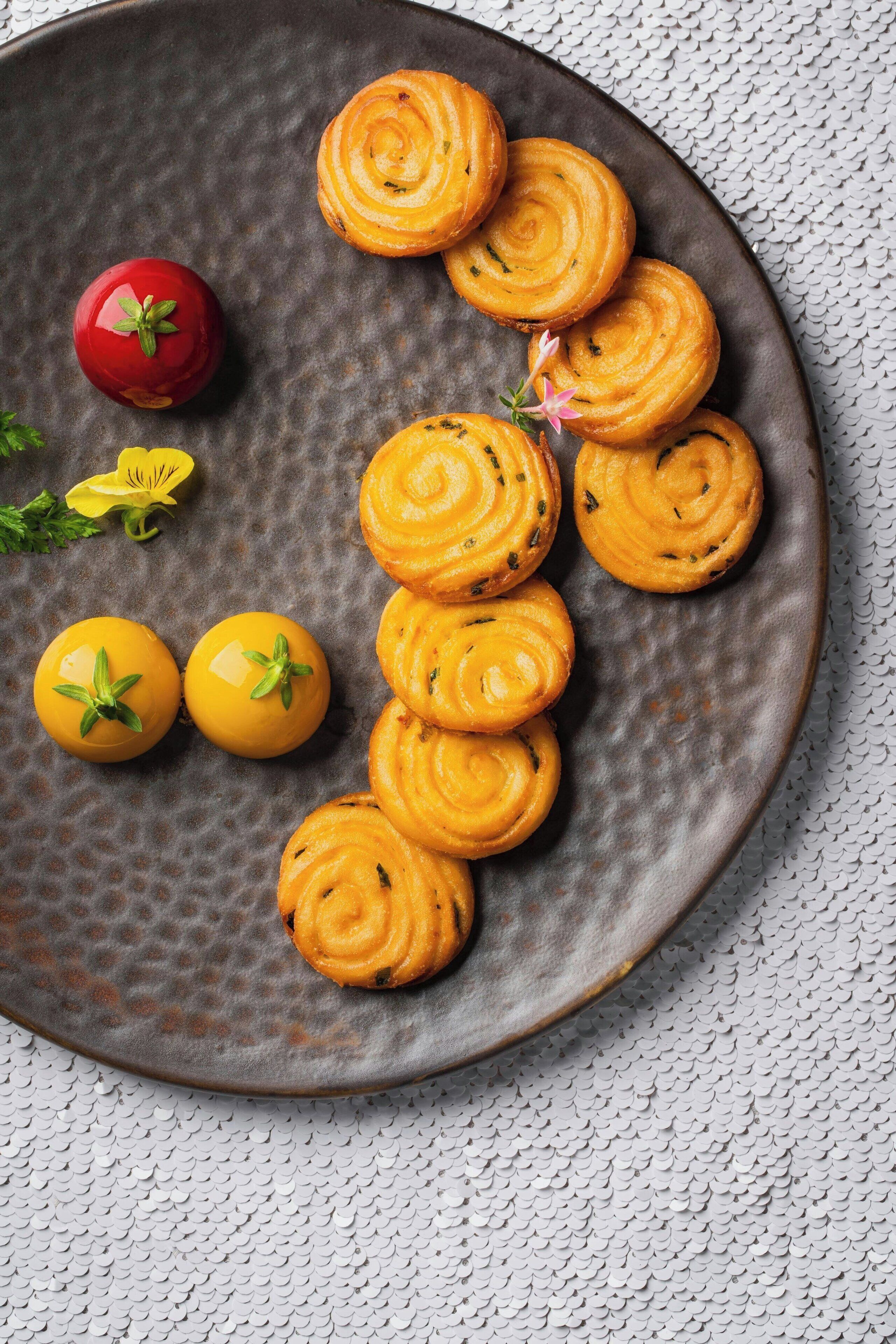 A plate with sliced, spiral-shaped food items, a red tomato, and two yellow tomatoes with green leaves.