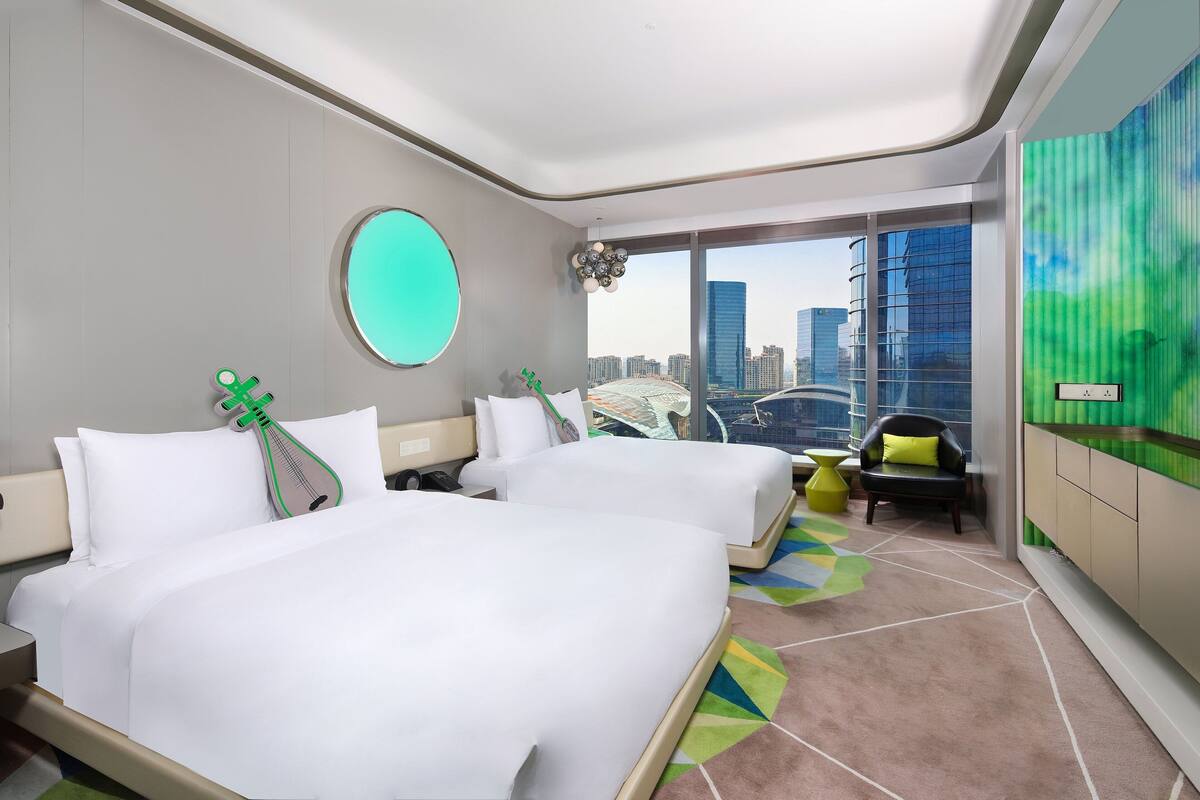 A modern hotel room with two beds, a large window with a city view, a round mirror, and a green decorative item on the wall.