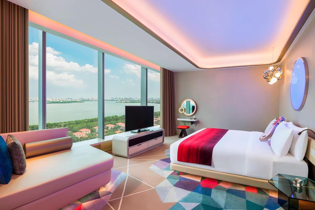 A modern hotel room with a large bed, a TV, a sofa, and a view of the city.