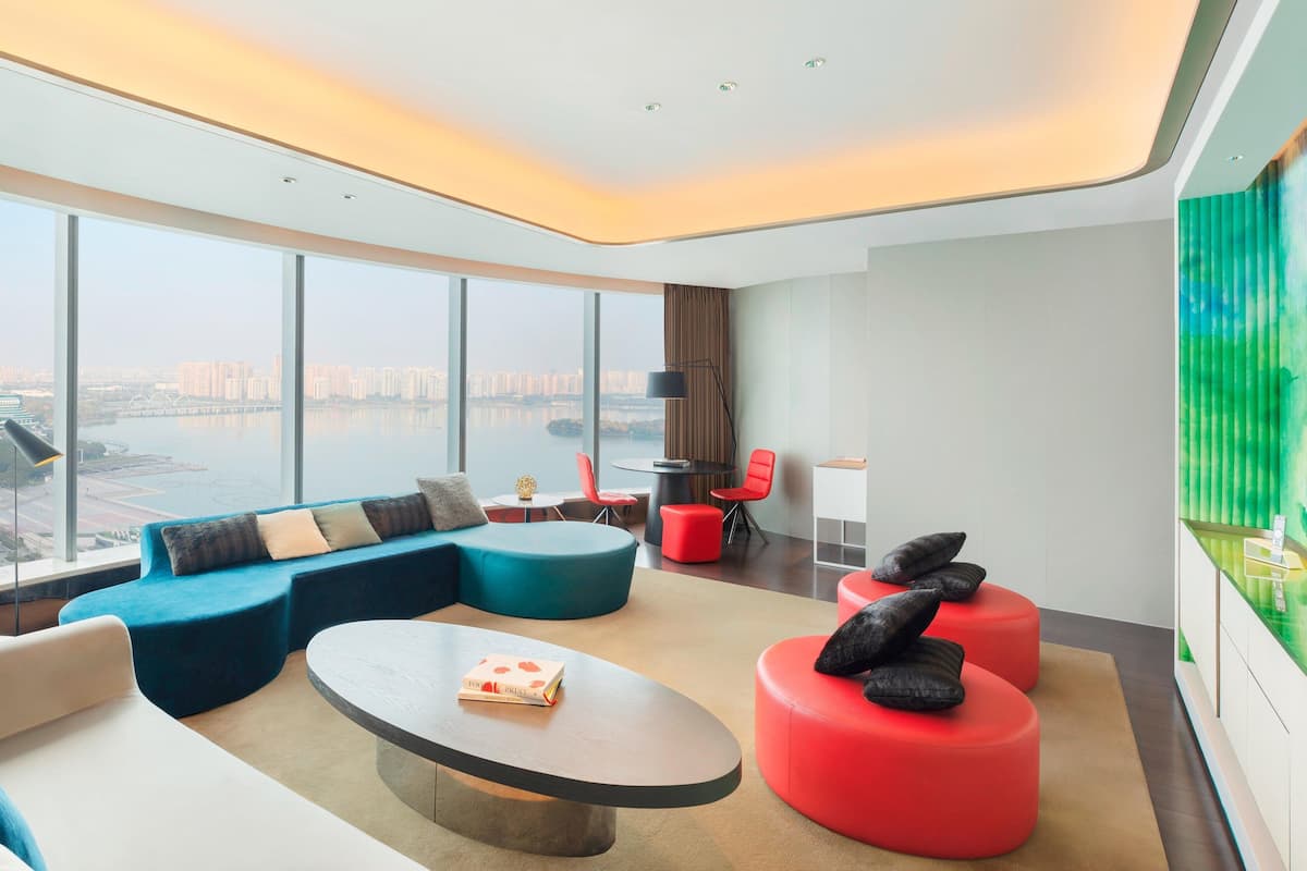 A modern hotel room with a large window offering a city view, a curved sofa, a round coffee table, and red ottomans.