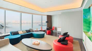 A modern hotel room with a large window offering a city view, a curved sofa, a round coffee table, and red ottomans.