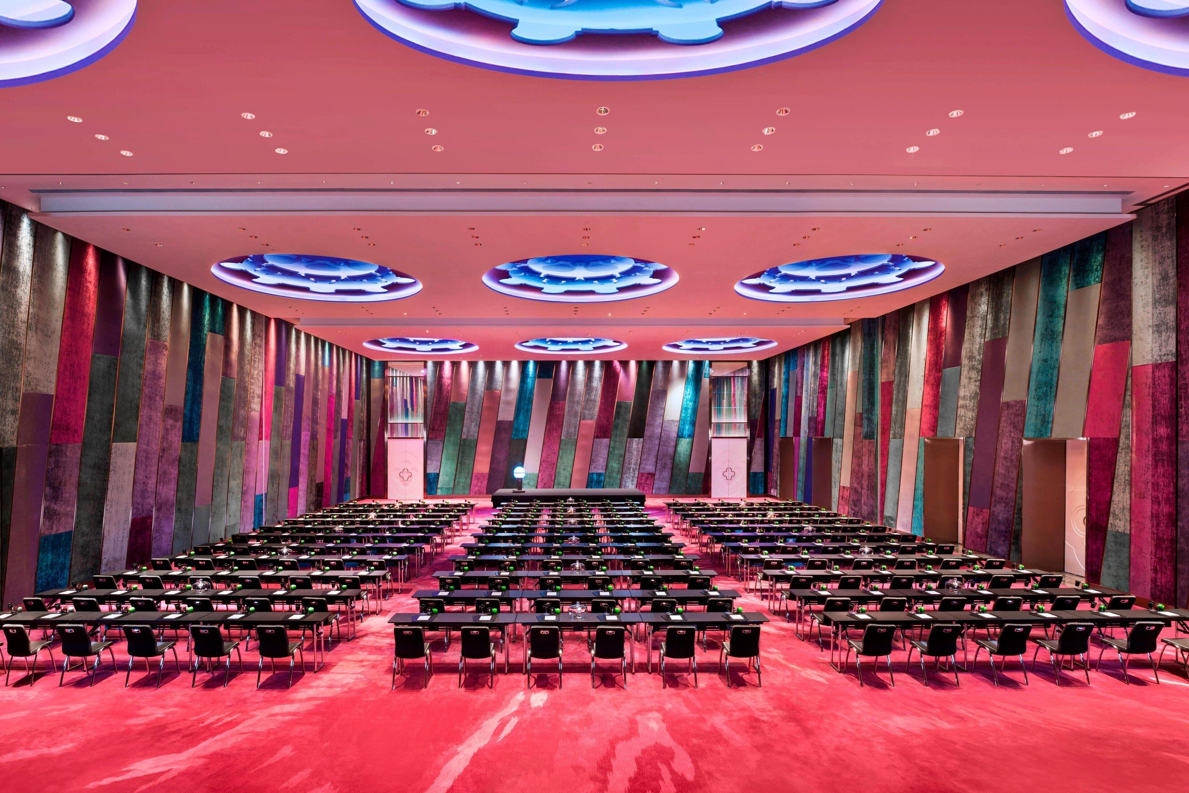 A conference room with rows of tables and chairs, a stage at the front, and a colorful, modern ceiling design.