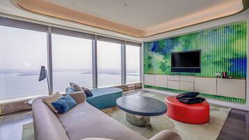 A modern living room with a large window, a sofa, a red ottoman, and a flat-screen TV mounted on a green wall.