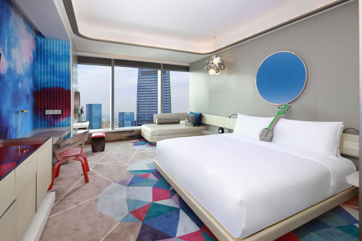 A modern hotel room with a large bed, a desk, a chair, a sofa, and a view of the cityscape.