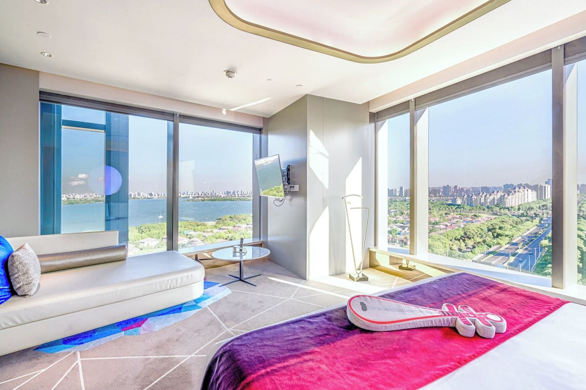 A modern hotel room with a large bed, a sofa, a small table, and a view of a cityscape and water.