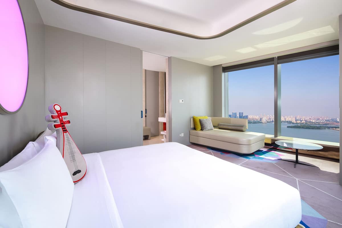 A modern hotel room with a large bed, a sofa, a small table, and a view of a cityscape through large windows.
