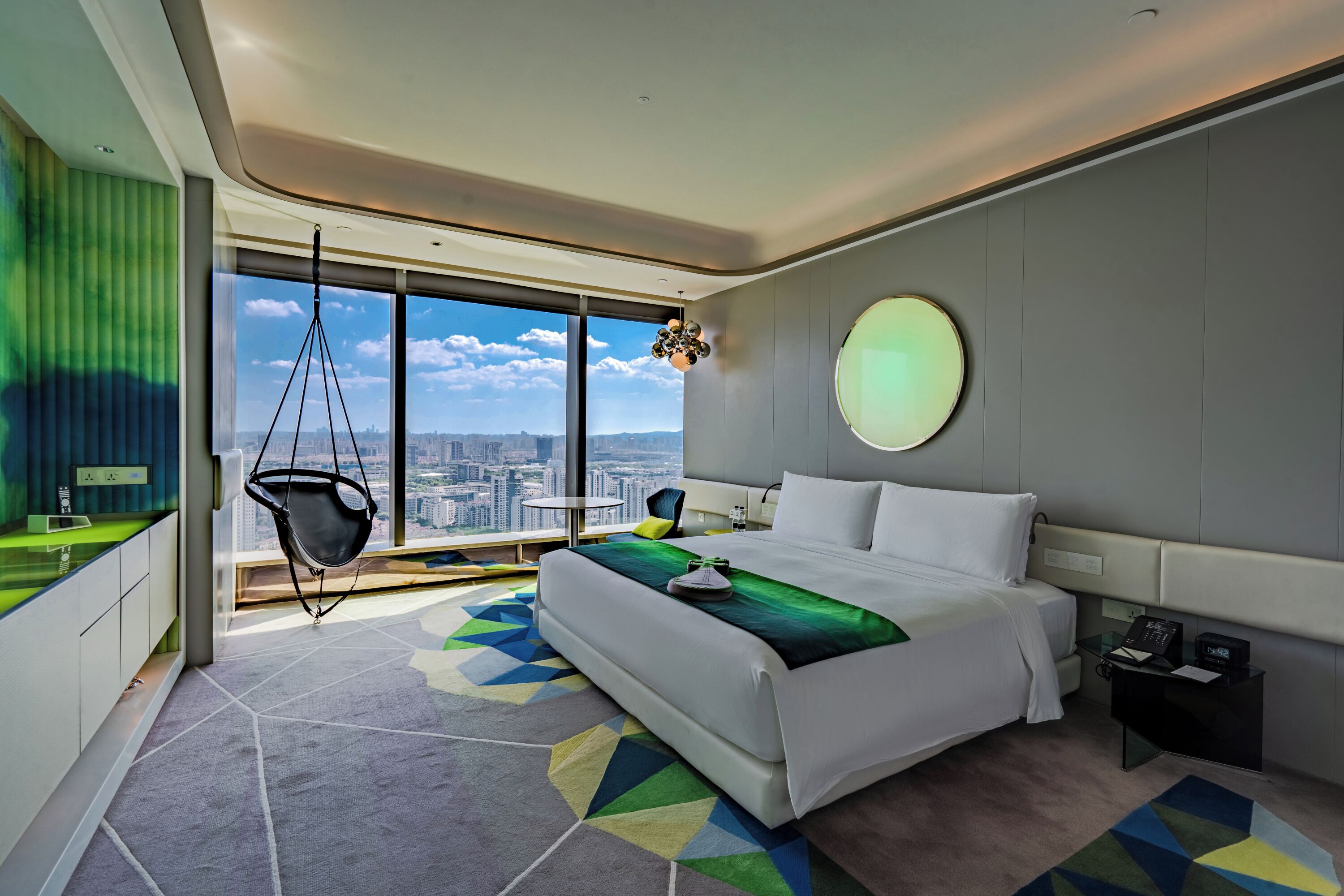 Camera d'albergo moderna con un letto grande, una sedia a dondolo, uno specchio rotondo e vista sulla città.