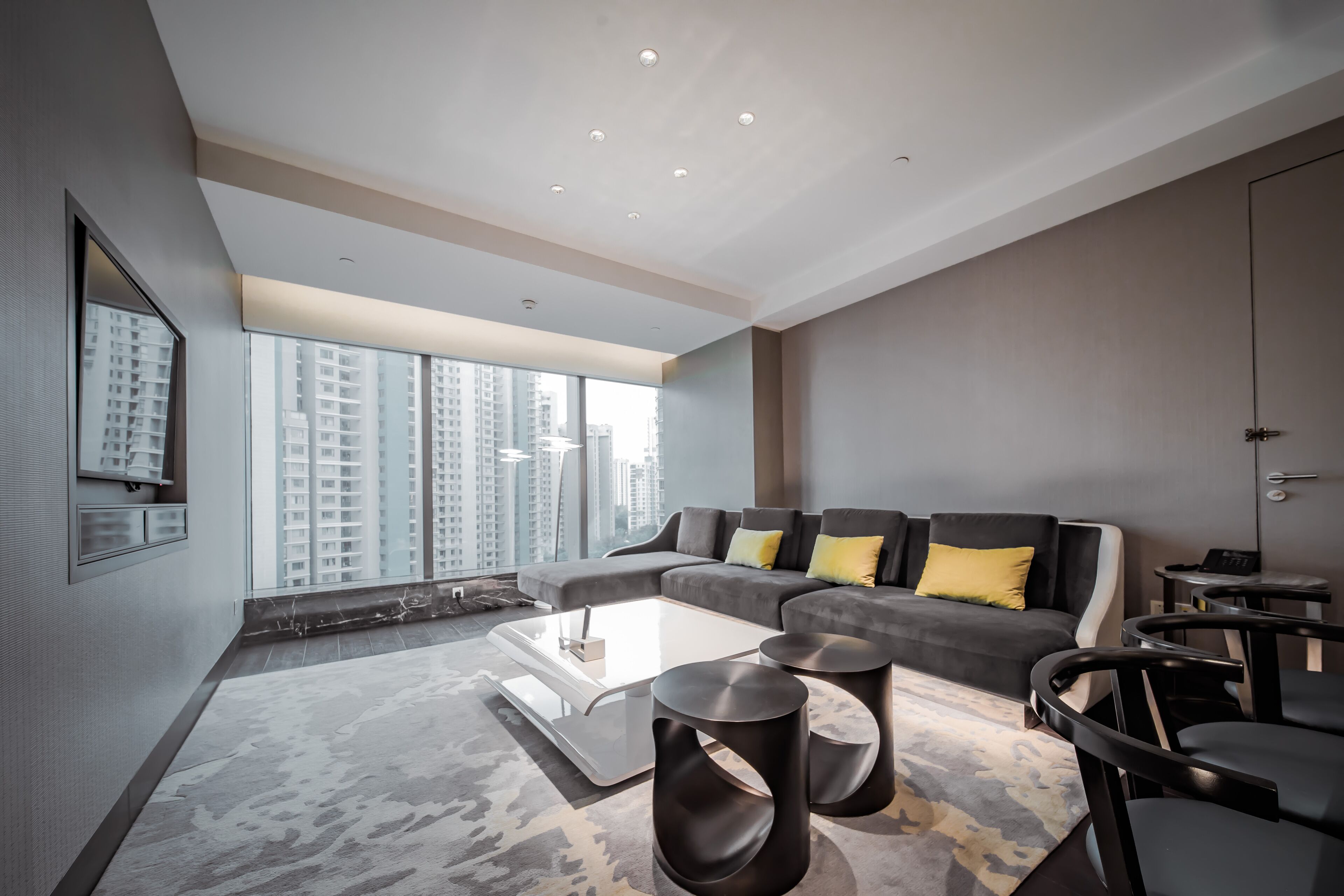 A modern living room with a grey sofa, black round tables, and a large window offering a city view.