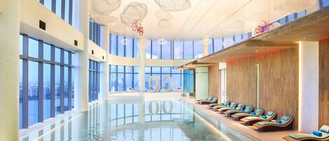 A modern indoor pool area with a clear view of the cityscape through large windows, a long pool with lounge chairs, and unique ceiling light fixtures.