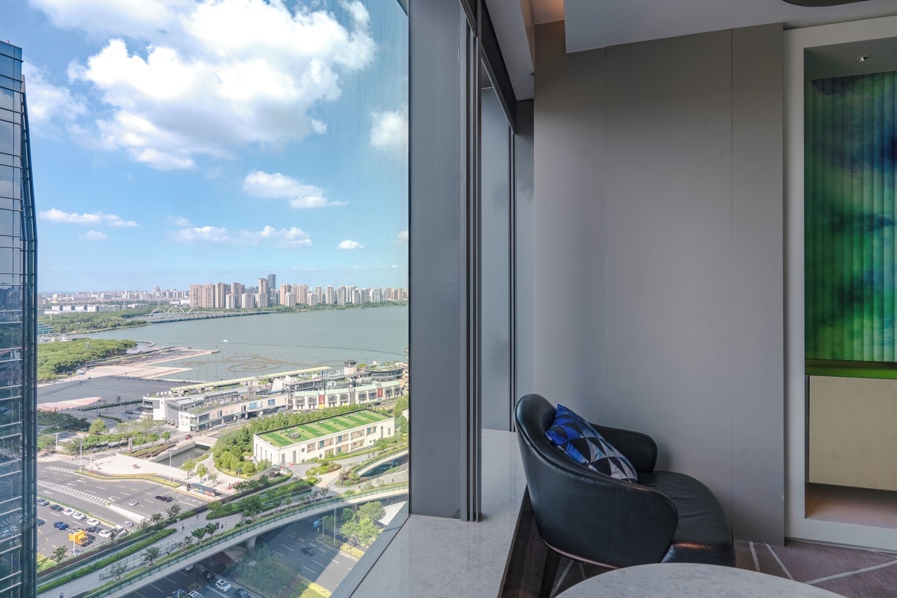 A modern hotel room with a large window offering a city view, a comfortable chair, and a small table.