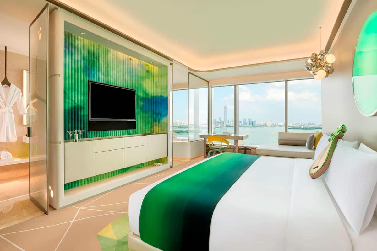 A modern hotel room with a large bed, a TV mounted on a green wall, a dining area with a view of the city, and a balcony with a cityscape view.