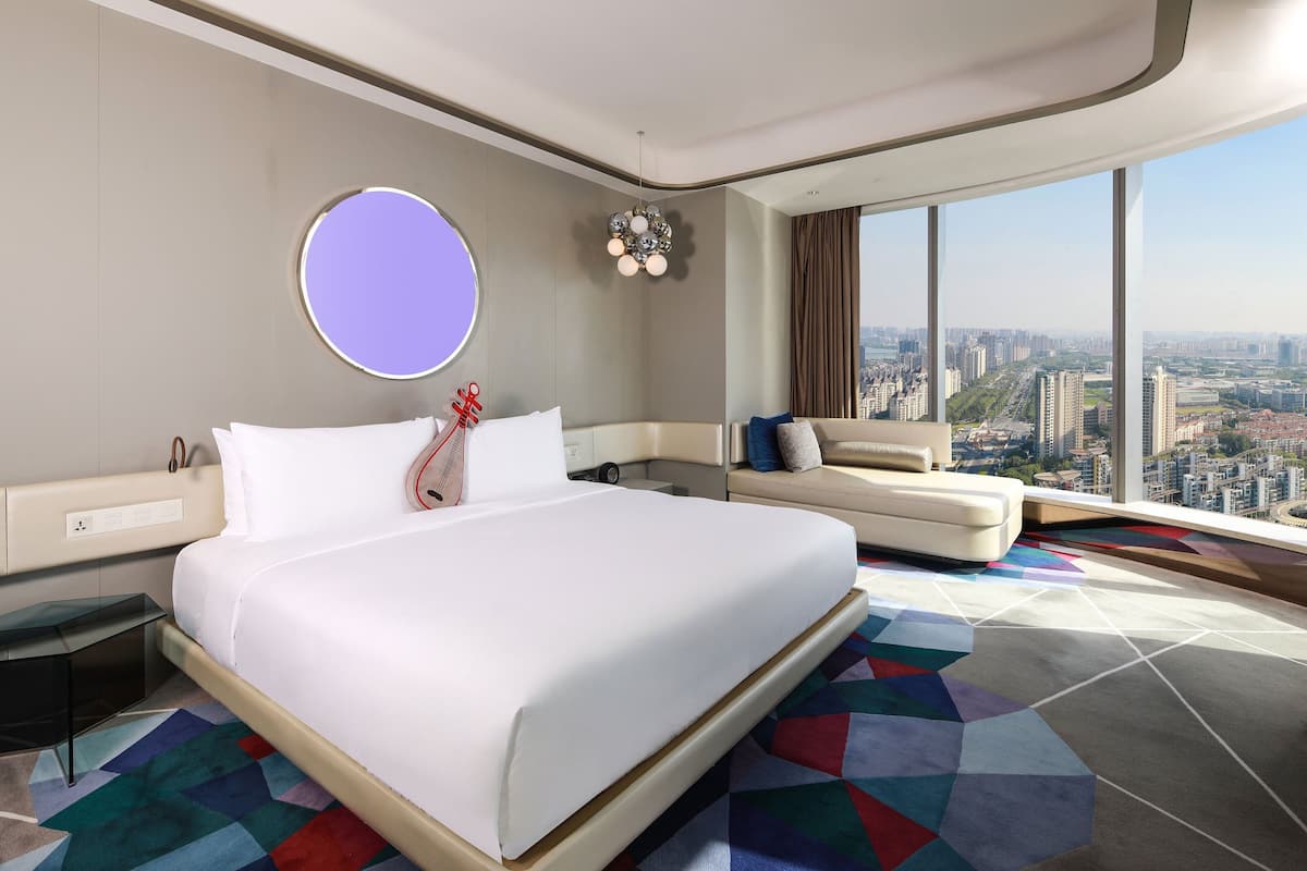A modern hotel room with a large bed, a sofa, a round mirror, and a city view.
