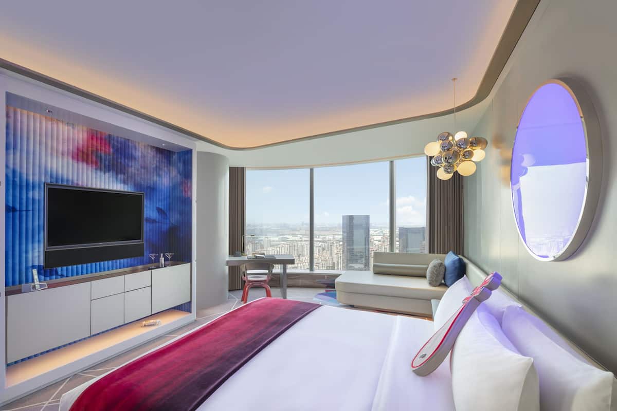A modern hotel room with a large bed, a TV mounted on a wall, a seating area with a view of the city, and a large window.