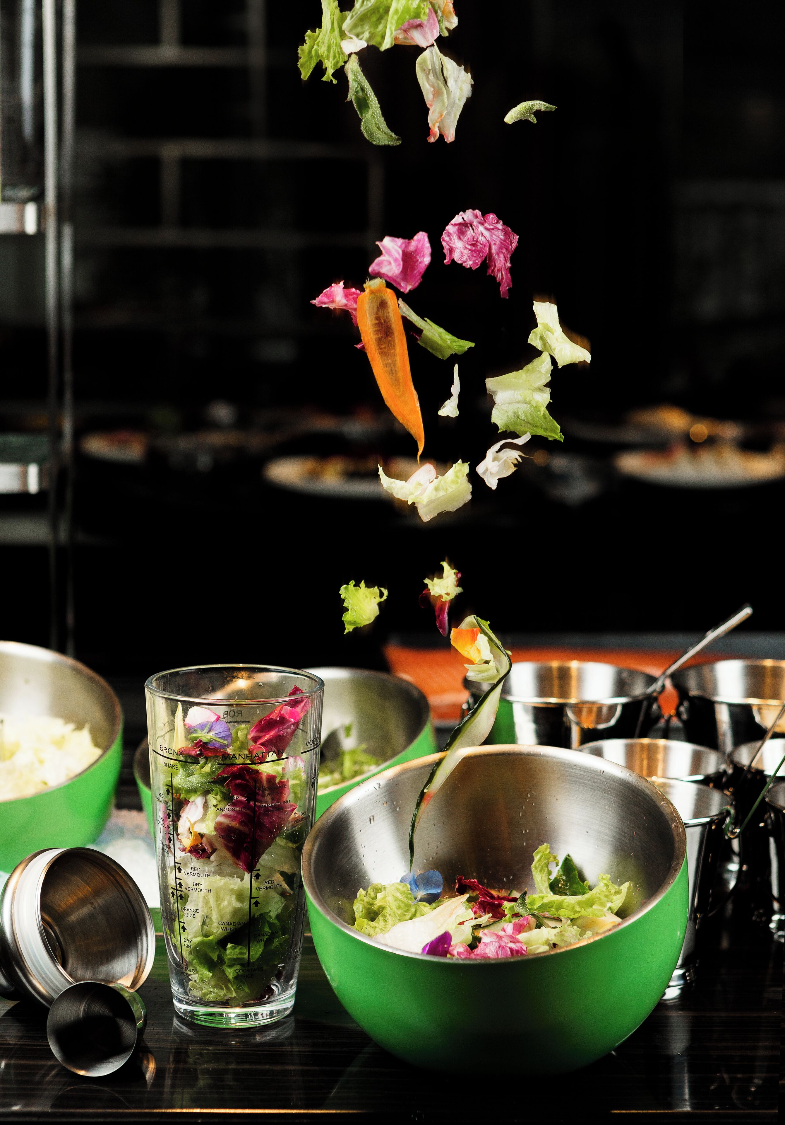 A dining table with a green bowl, a clear glass with a salad, and a metal bowl.