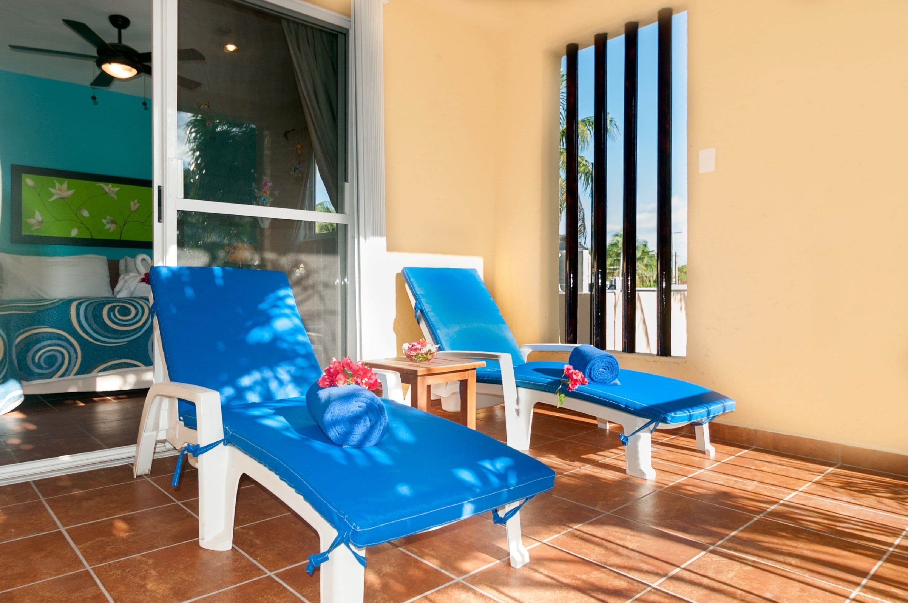 Una habitación iluminada por el sol, con dos sillones azules, una mesita con una flor y vistas a palmeras a través de una ventana.